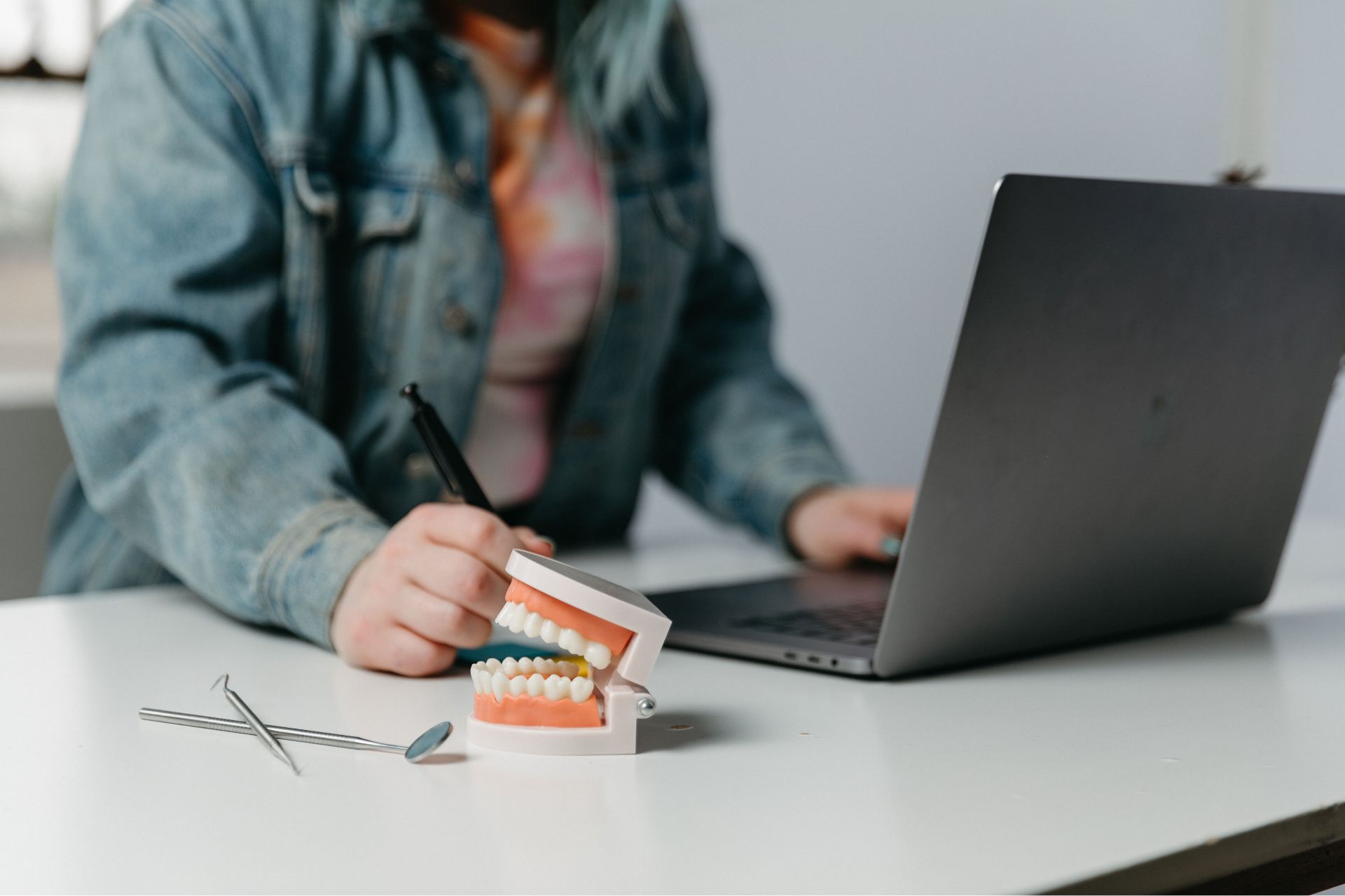 Dental model and tools on desk with person using laptop and pen in background.