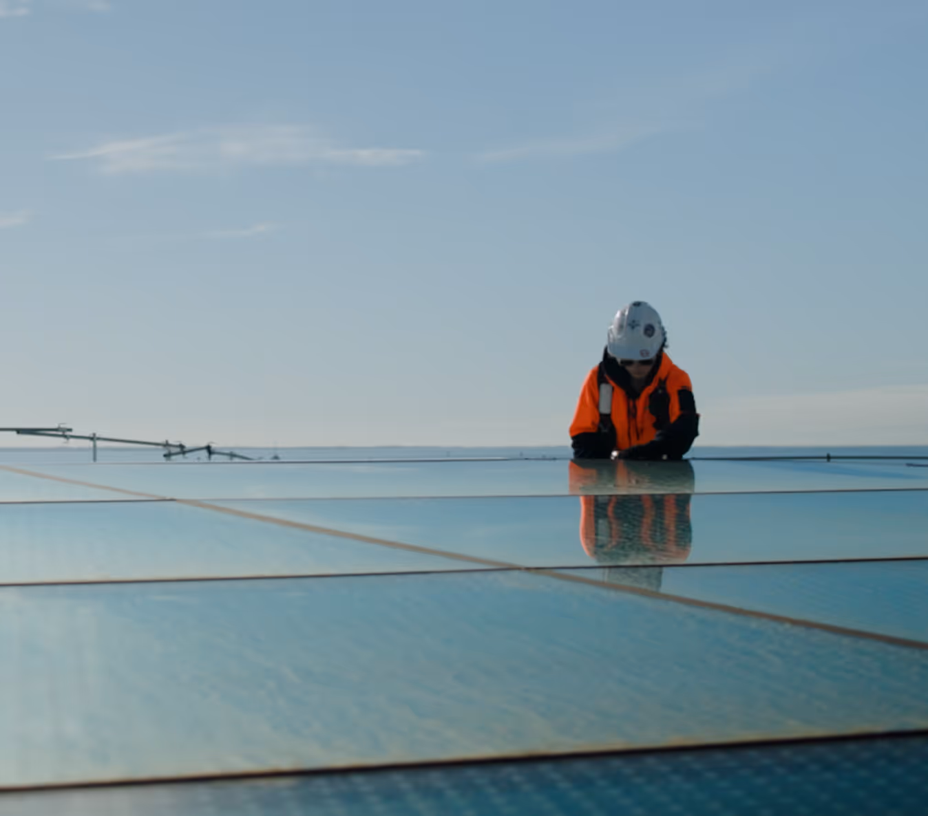 Worker in orange safety jacket and white helmet inspecting solar panels on a roof under clear blue sky.