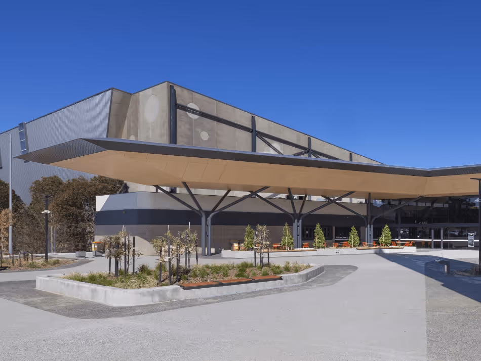 Modern building entrance with a large cantilevered roof, landscaped planters, and clear blue sky.