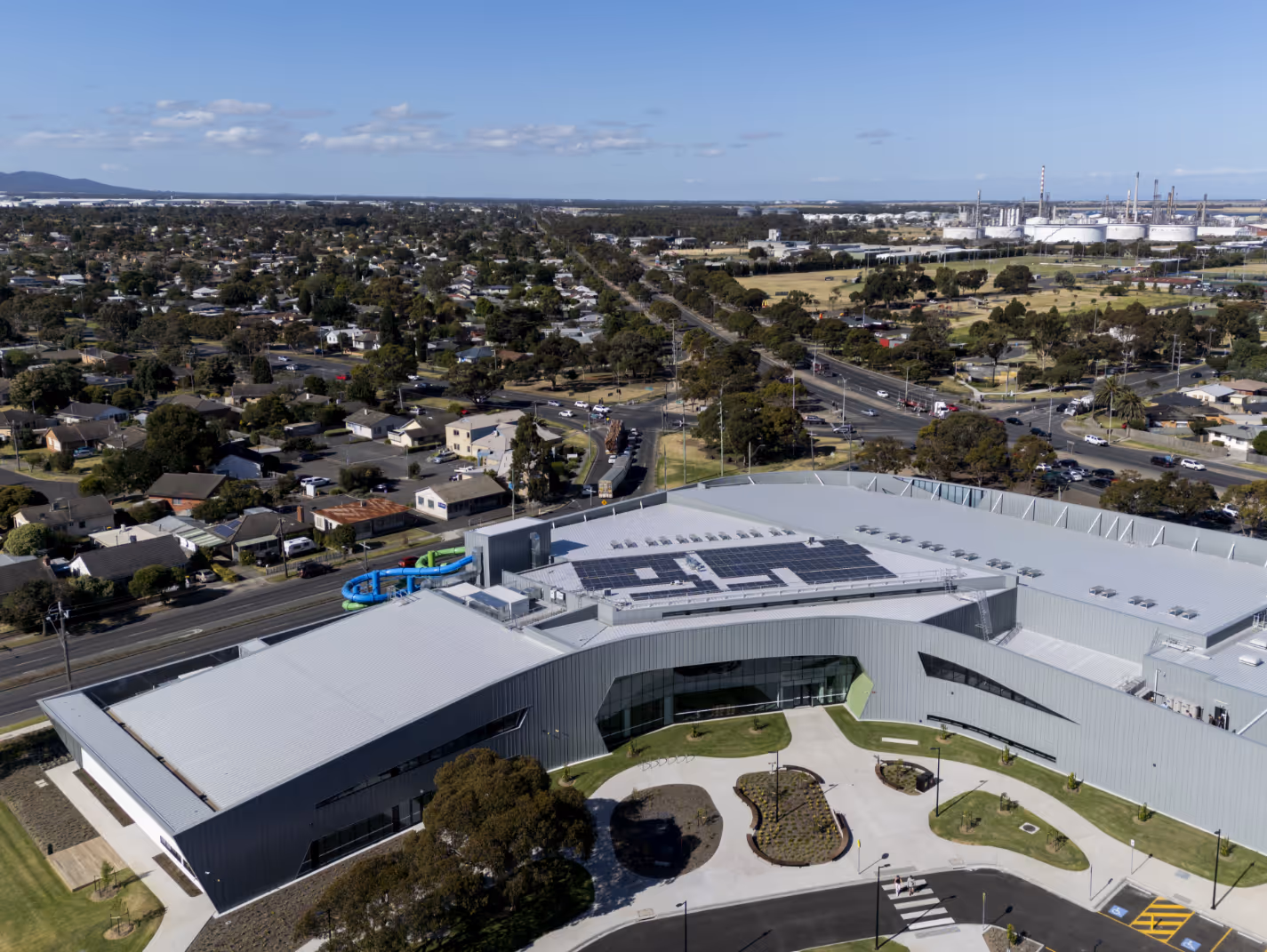 Aerial view of a large modern building with solar panels on the roof, surrounded by green lawns and residential neighborhood under a clear sky.