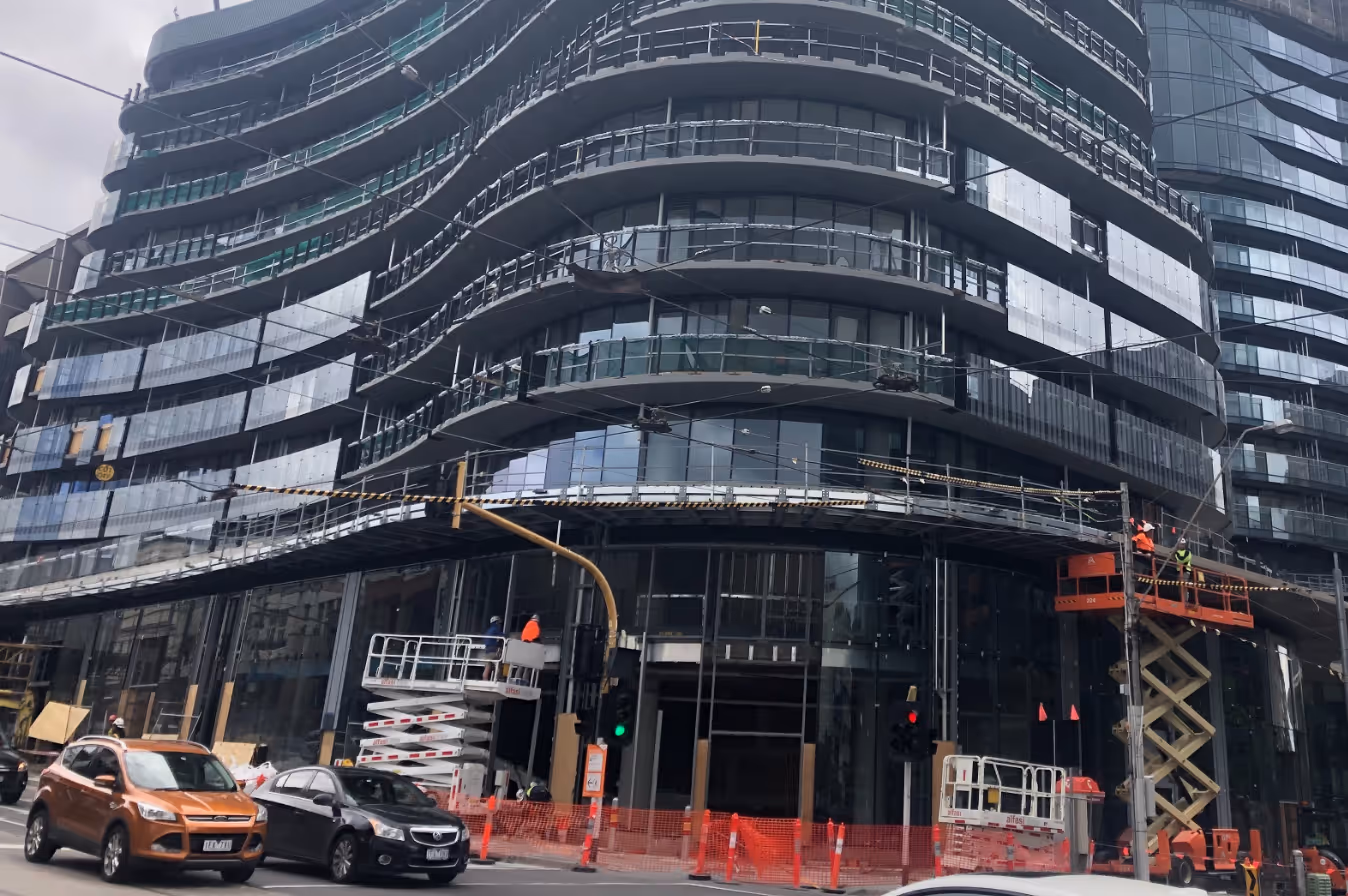 Modern curving glass building under construction with workers on lifts and cars on street below.