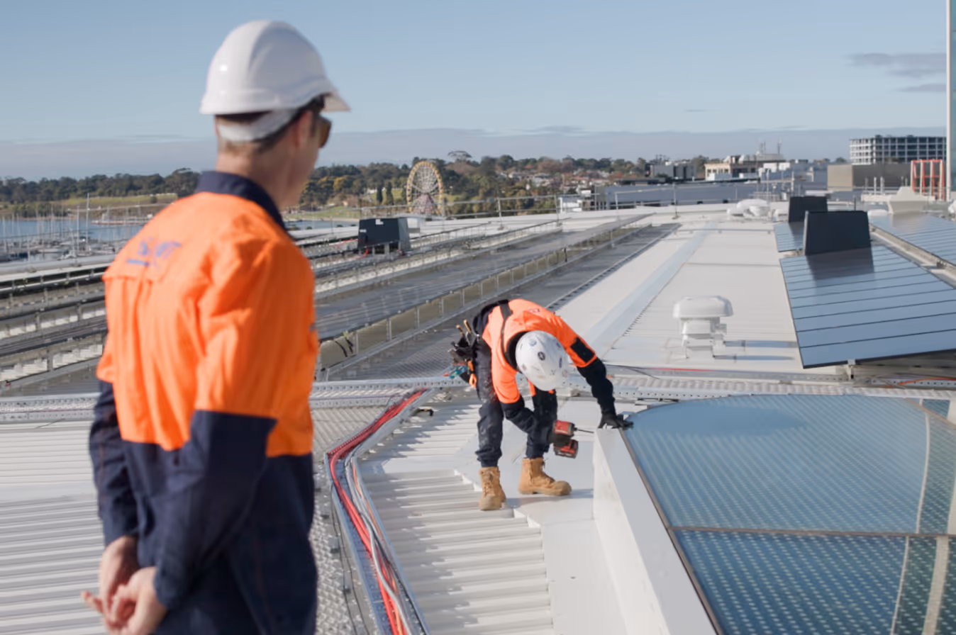 Two construction workers in orange and navy uniforms and white helmets working on a rooftop with solar panels and tools.