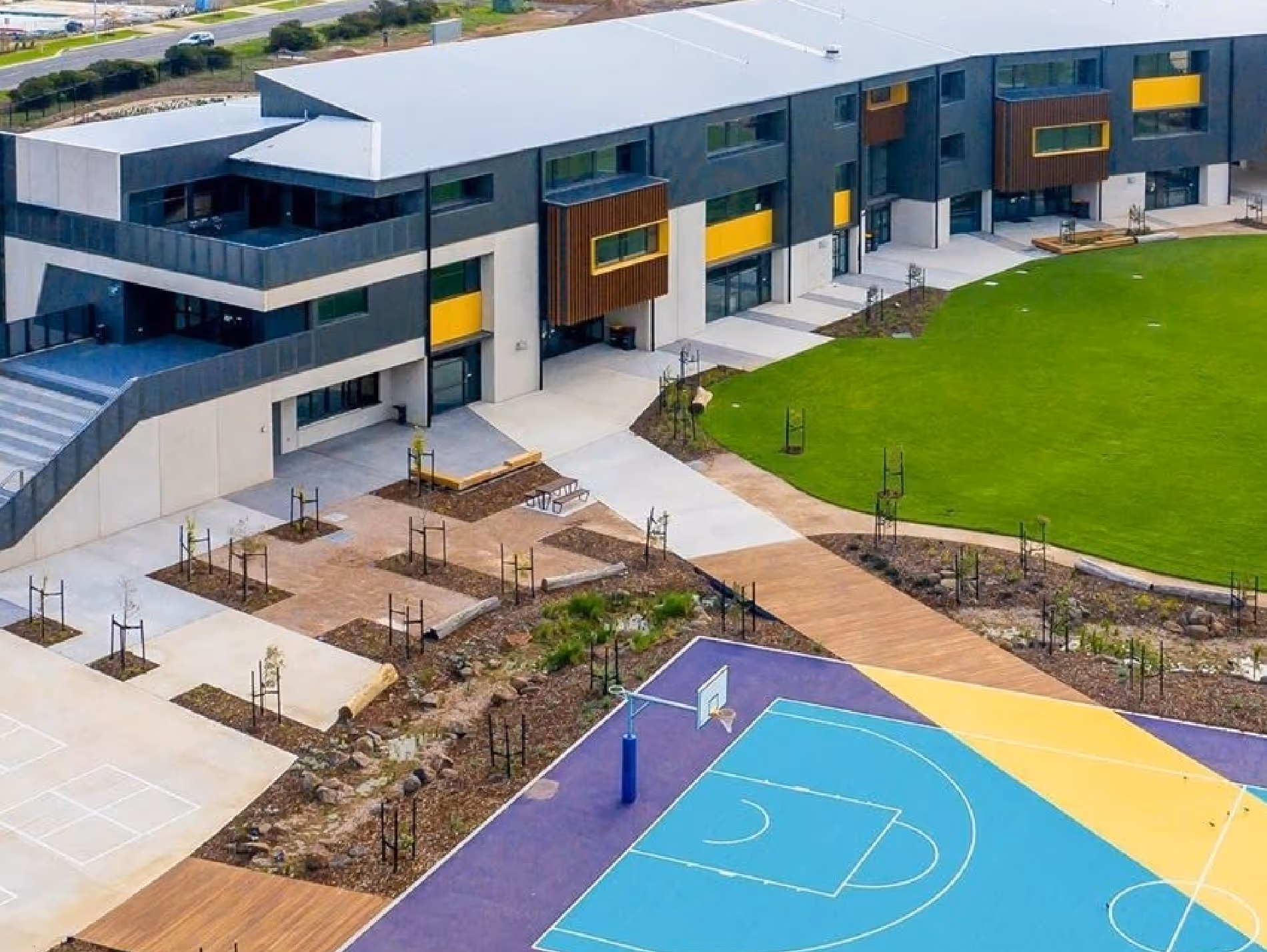Modern school building with yellow accents, green lawn, and a colorful basketball court with blue, yellow, and purple sections.