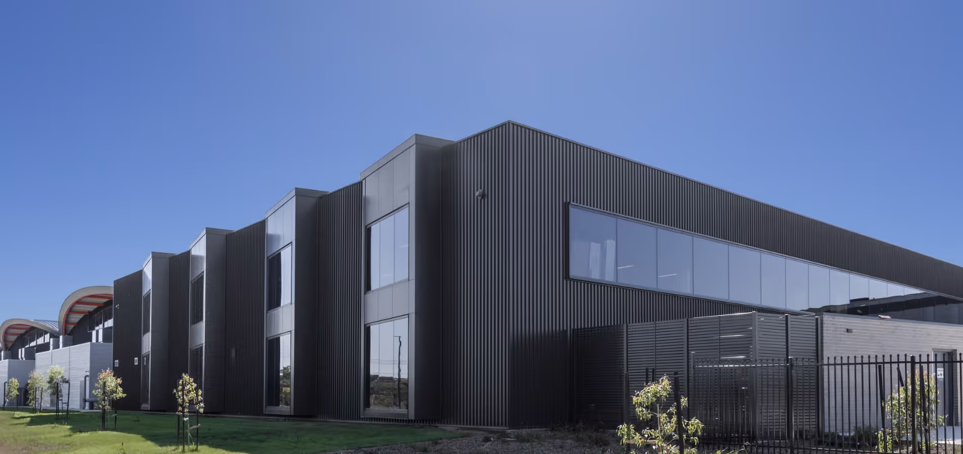 Modern industrial building with large windows and vertical black paneling under a clear blue sky.