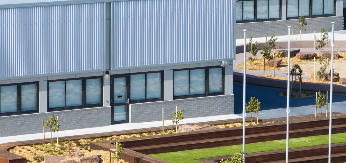 Modern building exterior with large windows, metal siding, wooden benches, young trees, and landscaped garden area.