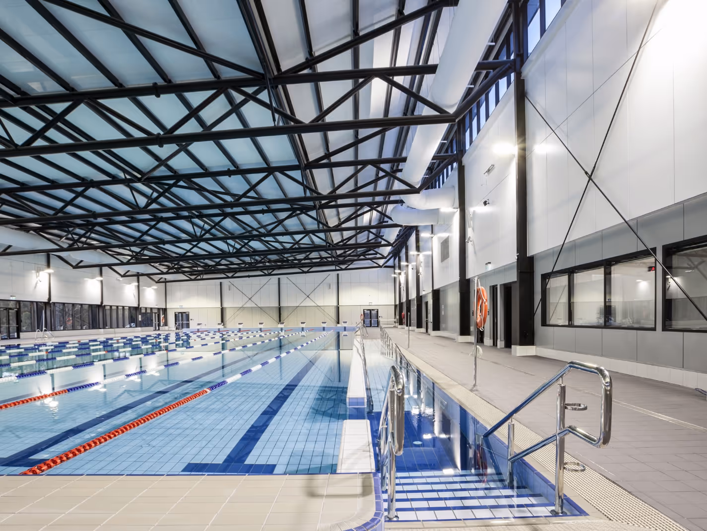 Empty indoor swimming pool with lane dividers, metal handrails, and large windows under a steel beam roof.