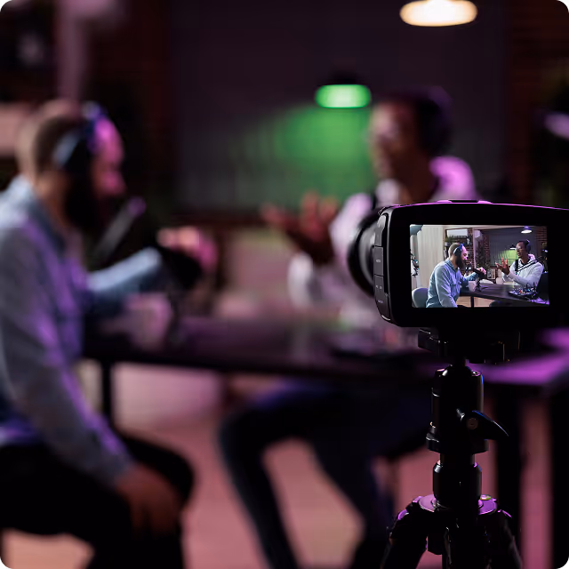 Two men wearing headphones recording a podcast at a table, viewed through the screen of a video camera on a tripod.