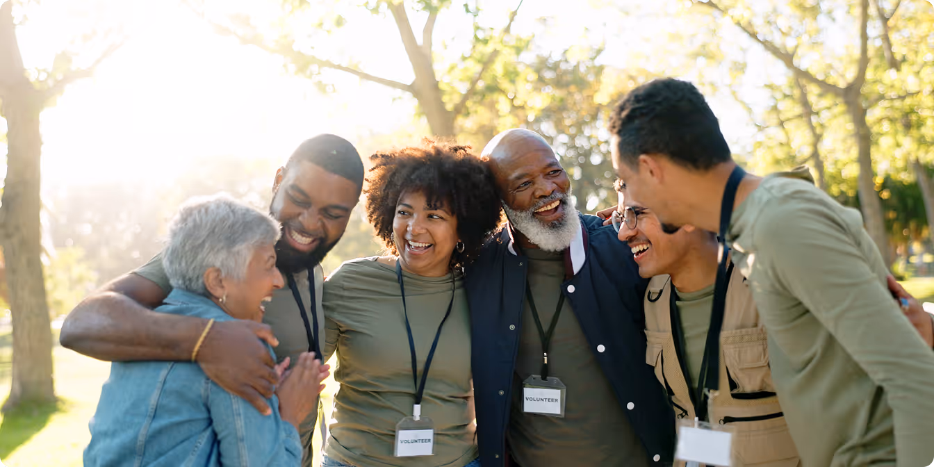Group of diverse volunteers smiling and embracing each other outdoors in a park on a sunny day.