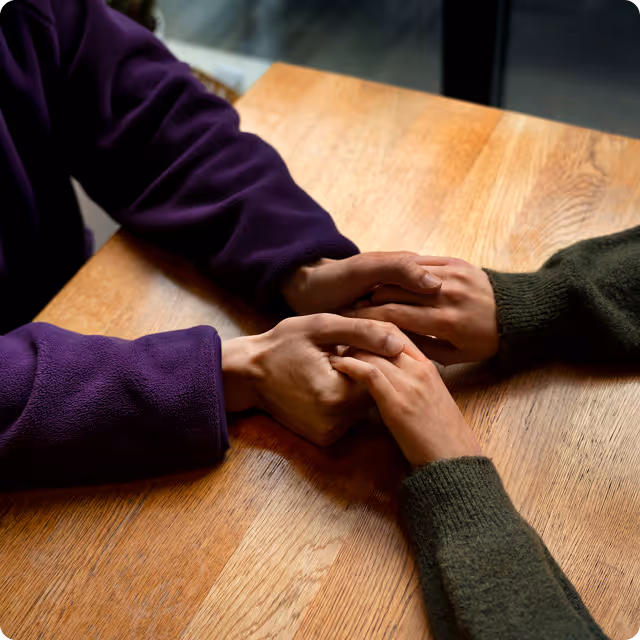 Two people sitting at a wooden table holding hands with a comforting gesture.