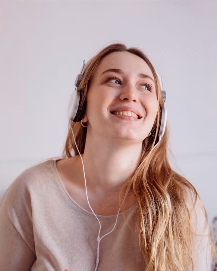 Smiling young woman with long blonde hair wearing white headphones and a beige top looking upward.