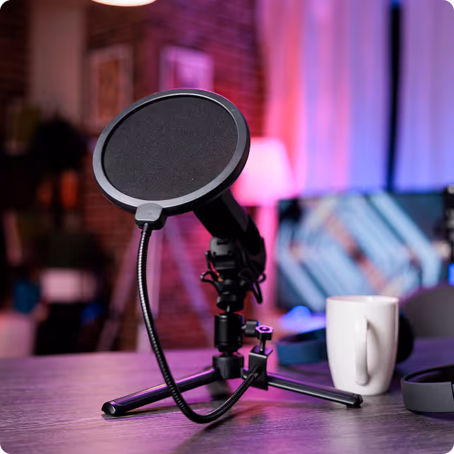 Close-up of a microphone with a pop filter mounted on a tripod stand on a table, with a white coffee mug and blurred colorful lights in the background.