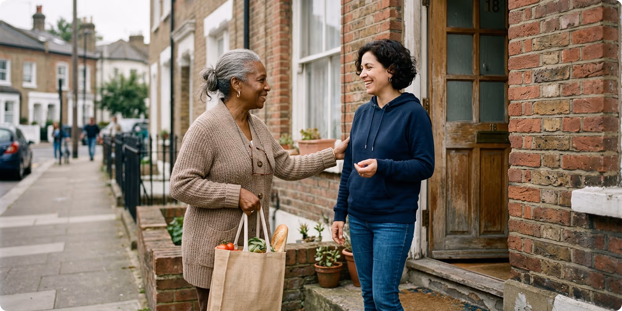 Two women smiling and conversing on a sidewalk outside a brick house, one holding a grocery bag filled with fresh produce.