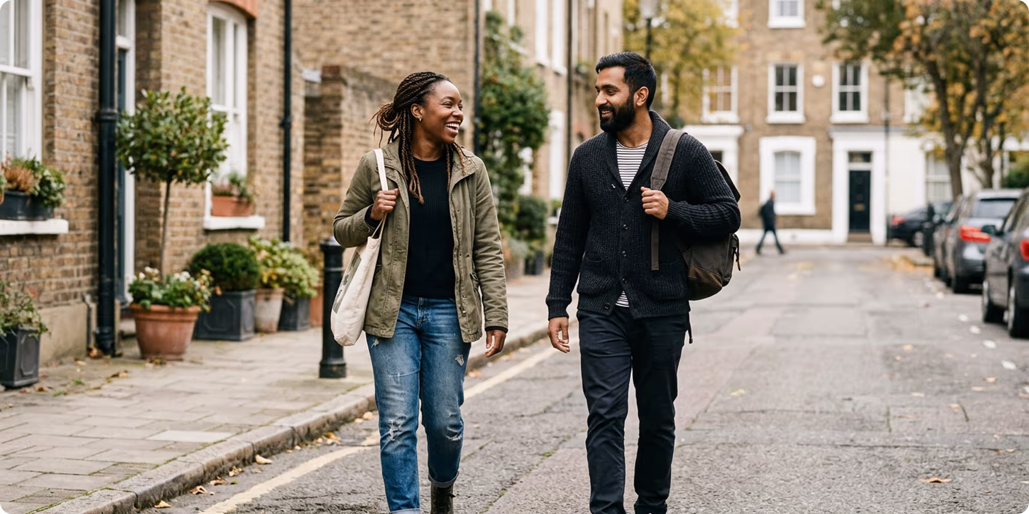 A smiling woman and a man walking and talking on a residential street with brick houses and potted plants.