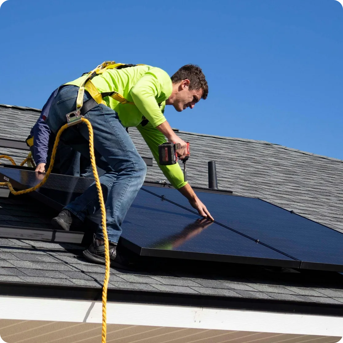 Worker in a safety harness installing solar panels on a rooftop under clear blue sky.