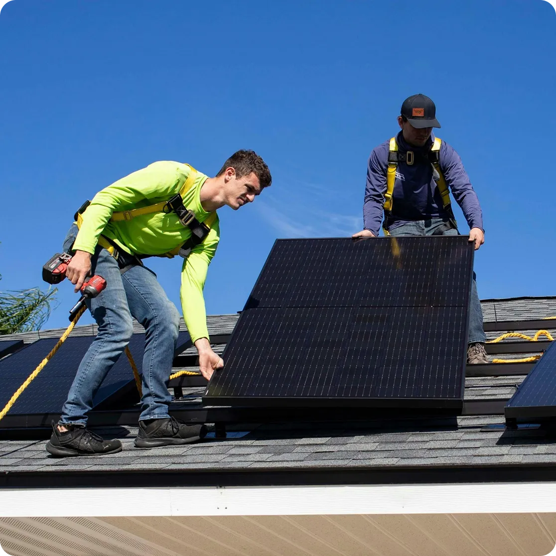 Two workers wearing safety harnesses installing solar panels on a rooftop under a clear blue sky.