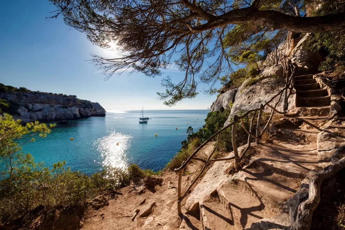 Pine-shaded stone steps descending to a sailboat anchored in a turquoise Menorcan cove