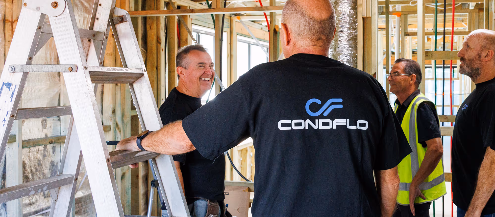 Four construction workers in a partially built interior with a ladder, one wearing a black shirt with the Condflo logo.