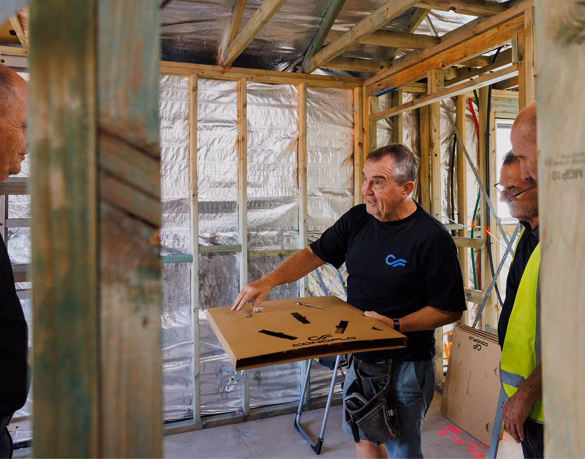 Construction worker holding a cardboard box with a diagram, explaining inside a building under construction with exposed wooden framing and insulation.