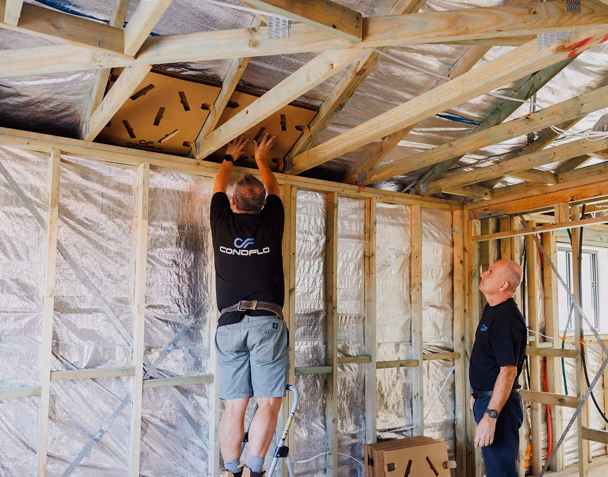 Two men installing a Condflo ventilation panel in the ceiling framework of a house under construction.