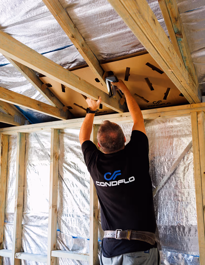 Man wearing a CONDFLO shirt installing a panel on wooden ceiling beams inside a building under construction.