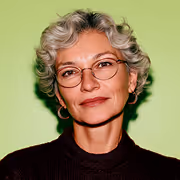 Portrait of a middle-aged woman with short gray hair, glasses, and a dark top against a green background.