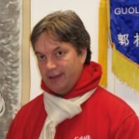 Mann mit braunen Haaren trägt einen roten Pullover und einen weißen Schal, steht vor einer Wand und einem blauen Banner.