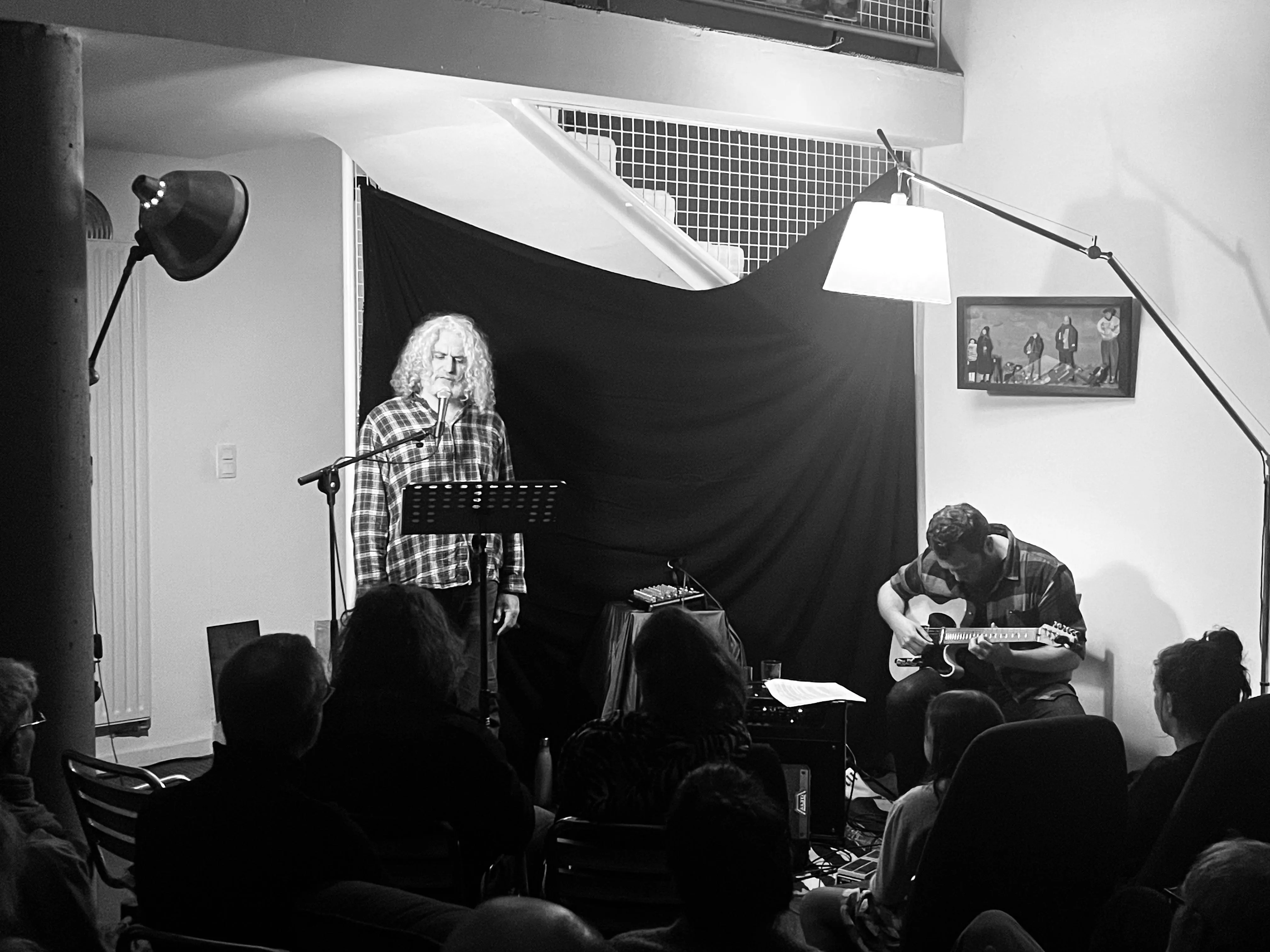 Black and white photo of a man singing into a microphone and another playing guitar in front of an audience in a small indoor venue.