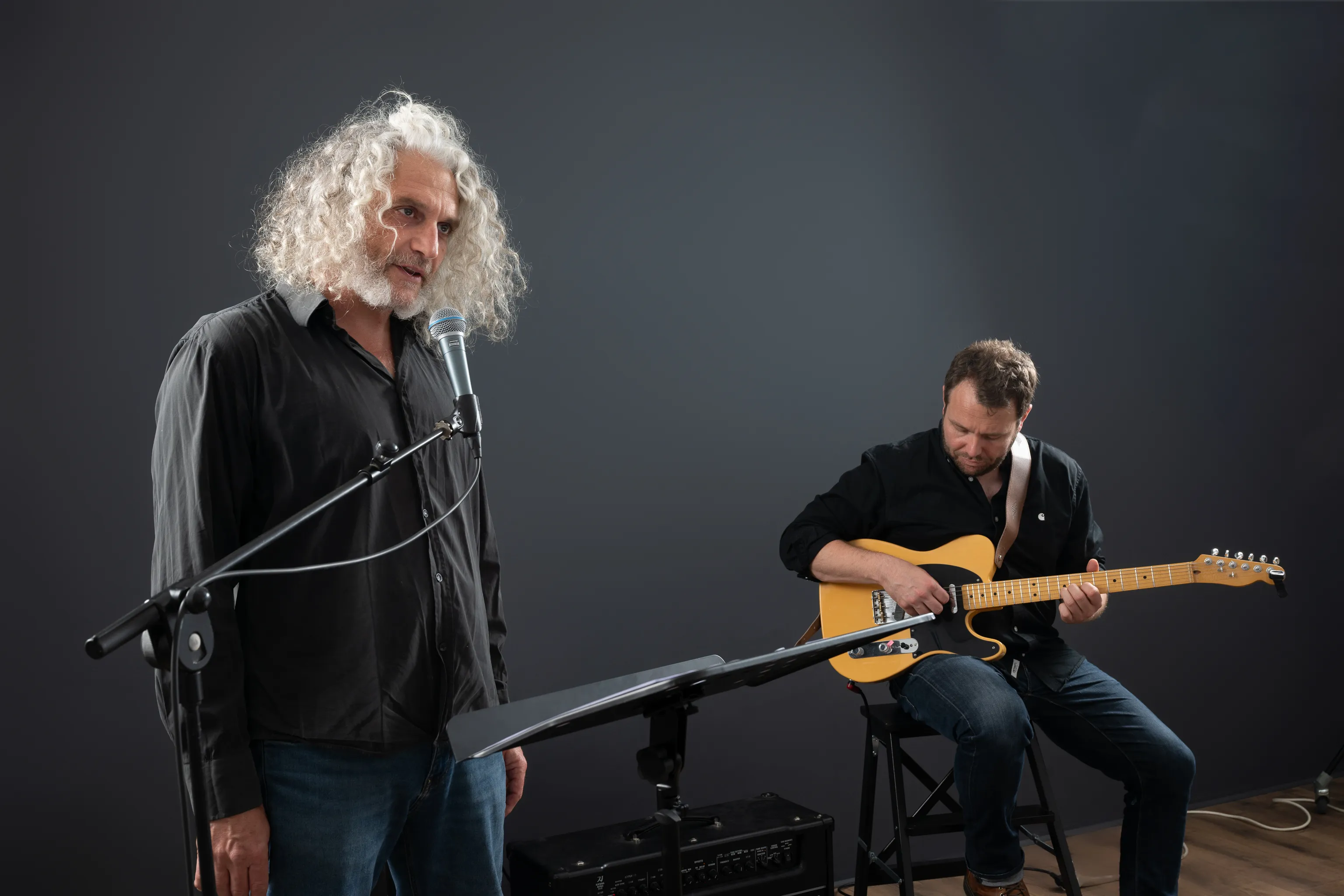Deux hommes en chemises noires pratiquent de la musique en studio, l'un chantant au micro et l'autre jouant de la guitare électrique jaune.