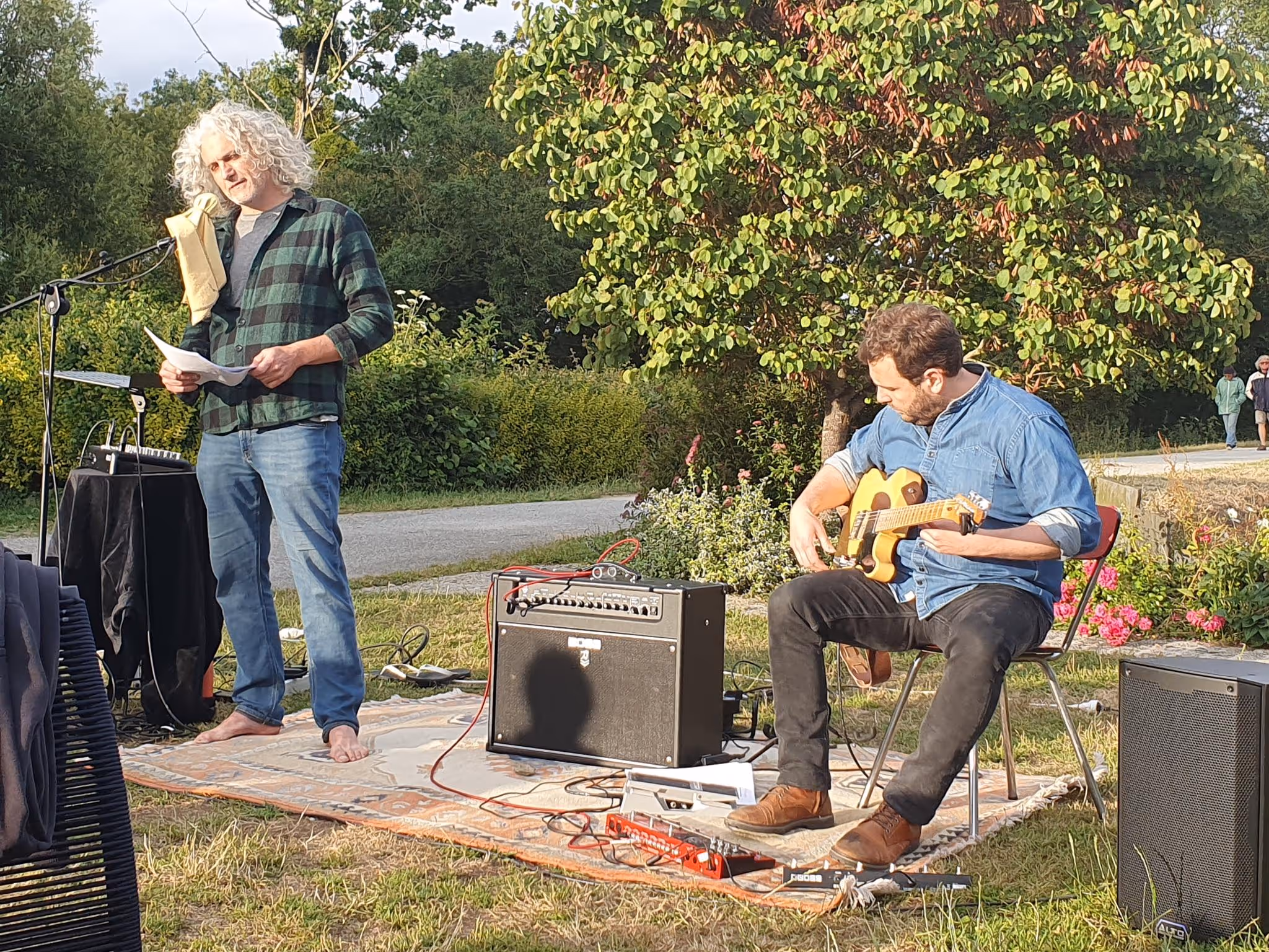 Un homme aux cheveux gris bouclés lit un texte devant un micro tandis qu'un autre homme joue de la guitare assis sur une chaise dans un jardin.
