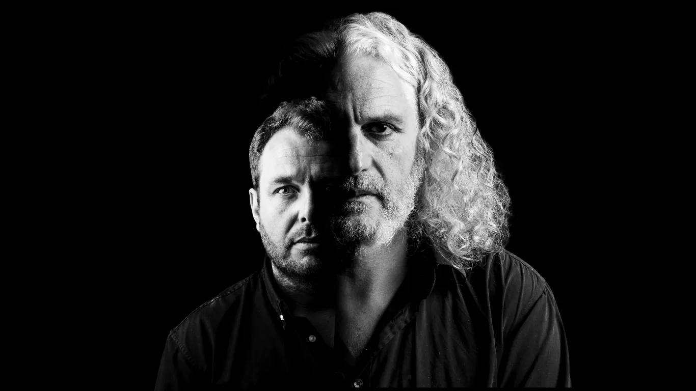 Black and white double exposure portrait of two men, one with curly white hair and beard and the other with short hair, against a black background.