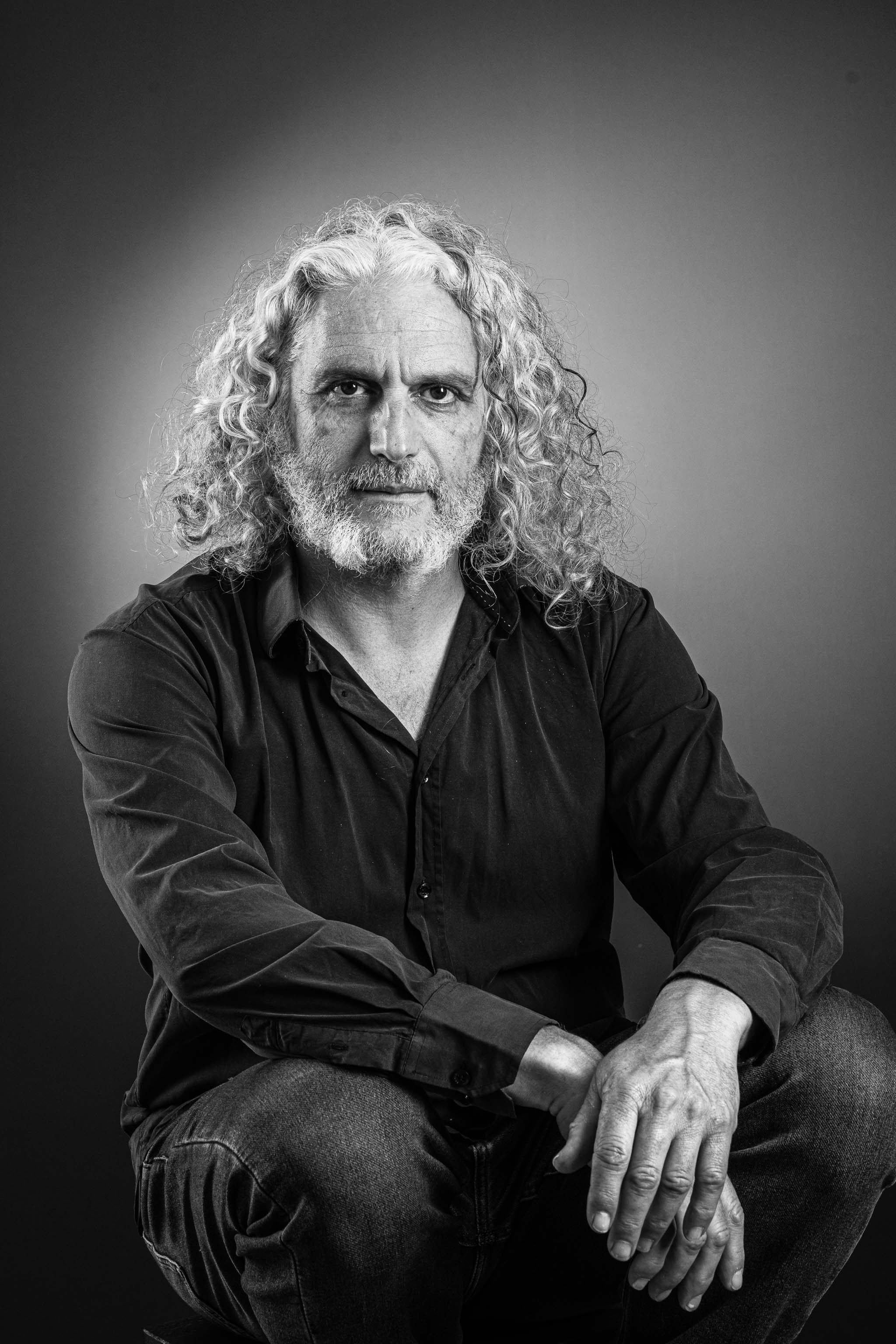 Portrait en noir et blanc d’un homme aux cheveux bouclés longs et à la barbe, en chemise et jean, assis.