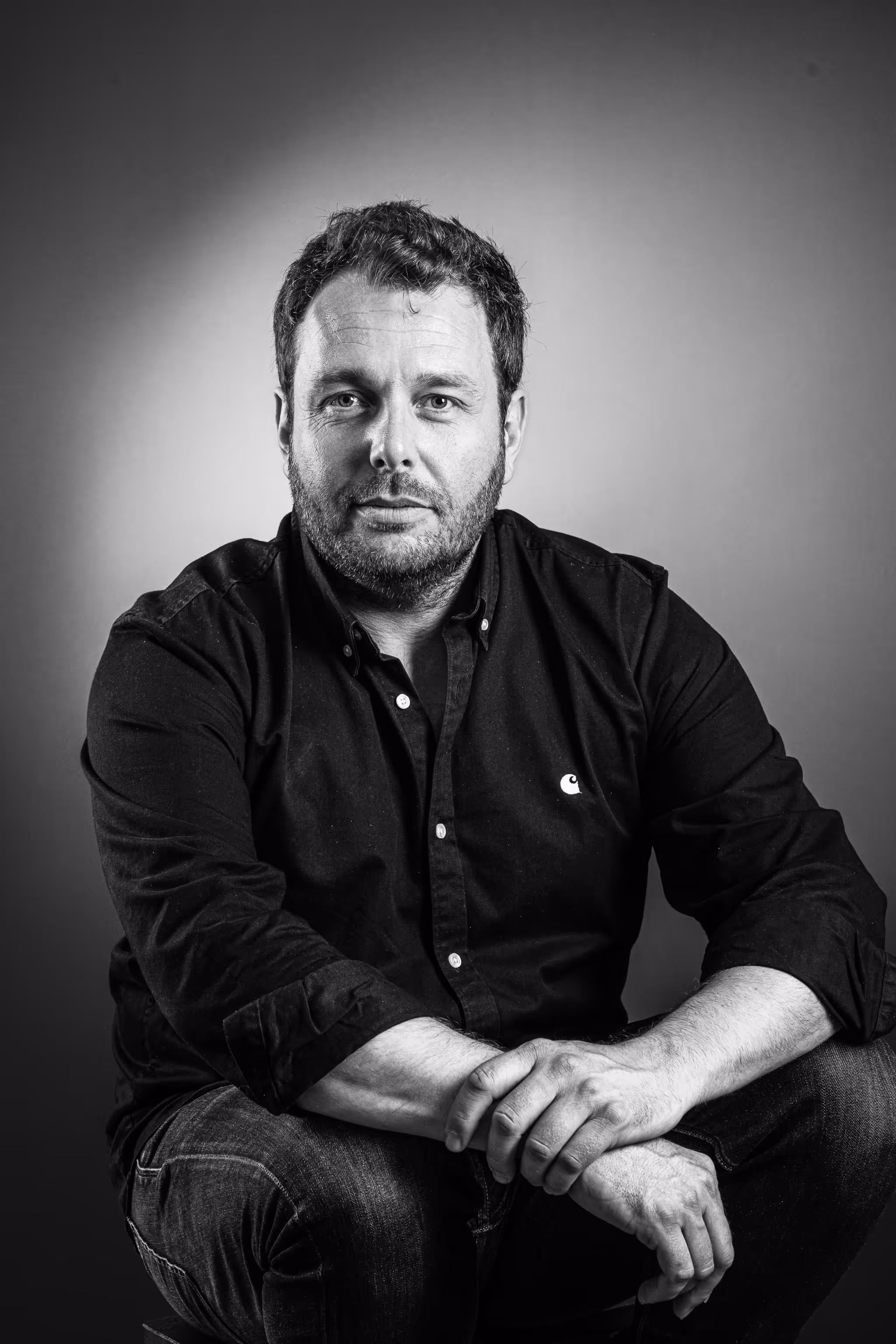 Portrait en noir et blanc d'un homme assis, regardant la caméra, portant une chemise noire.