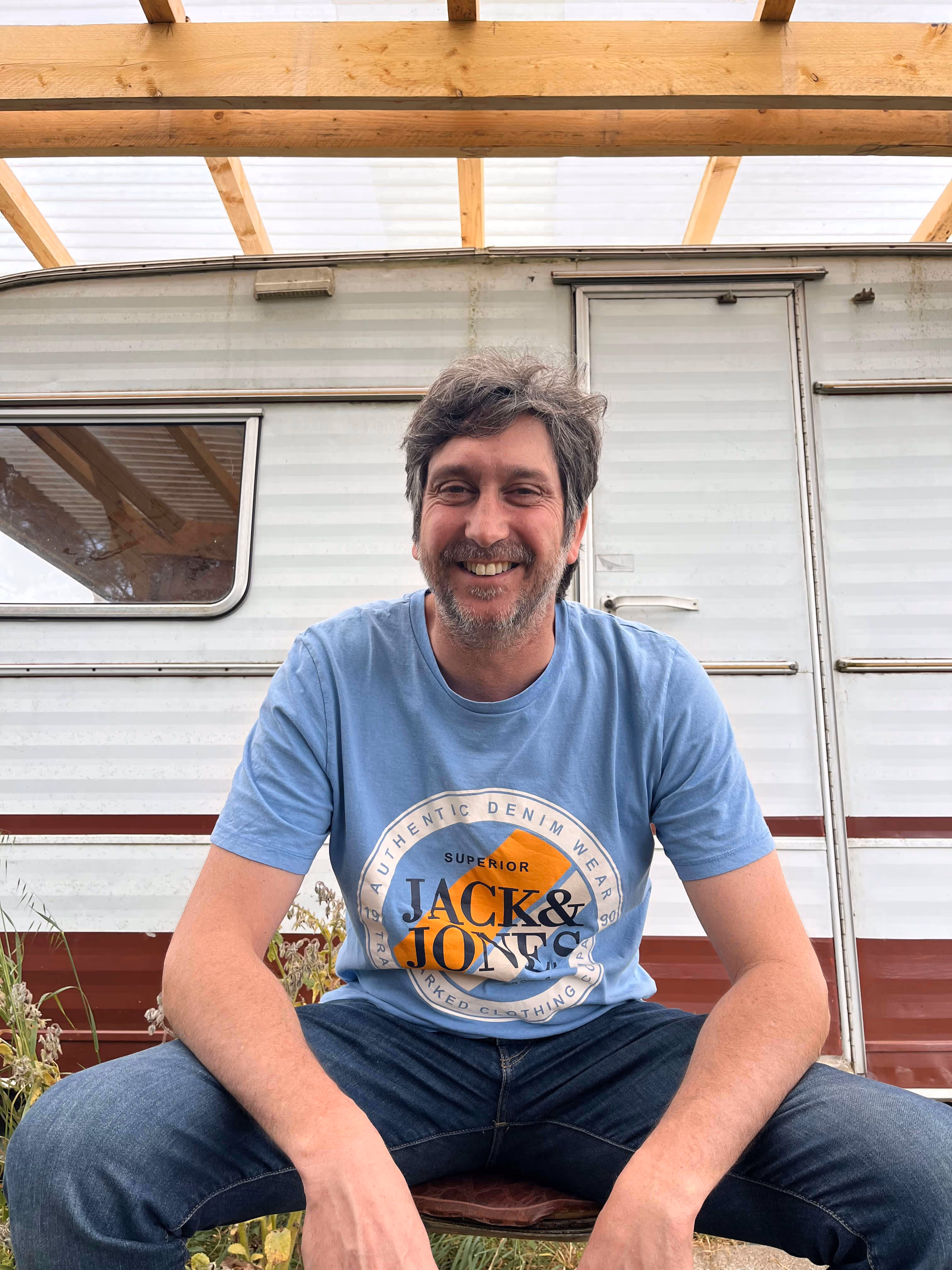 Homme souriant portant un t-shirt bleu Jack & Jones, assis devant une caravane sous une structure en bois.