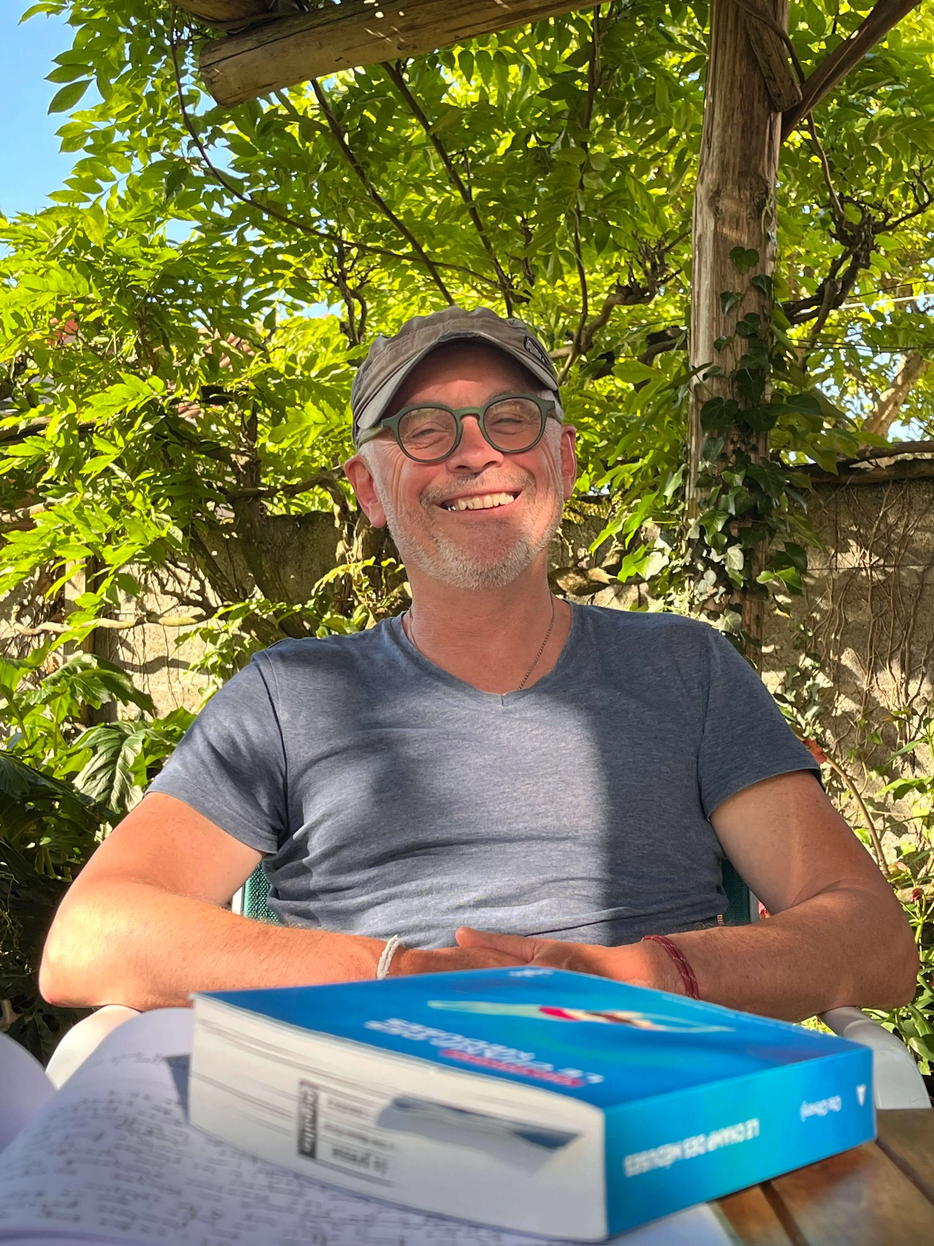 Homme souriant portant une casquette et des lunettes assis dehors avec un livre sur une table devant lui.