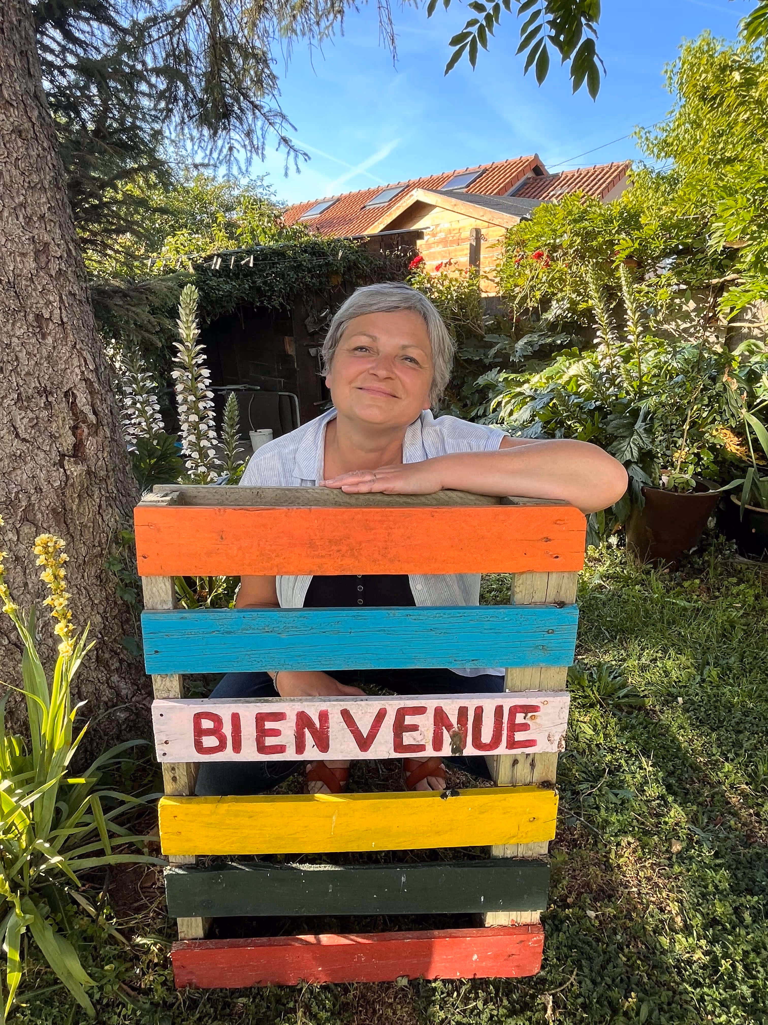 Femme souriante derrière un panneau coloré en bois avec le mot Bienvenue dans un jardin ensoleillé.
