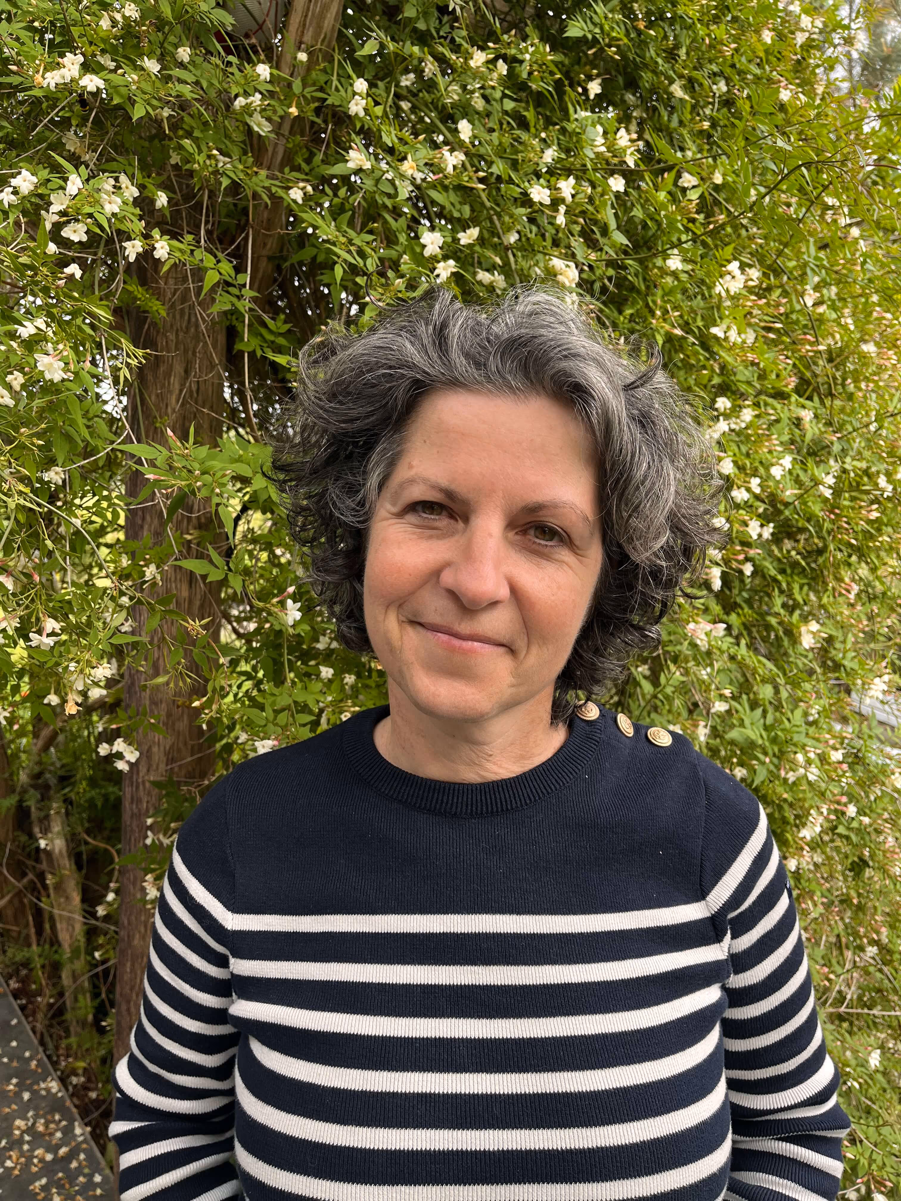Femme aux cheveux gris bouclés portant un pull à rayures noires et blanches devant un arbre fleuri.