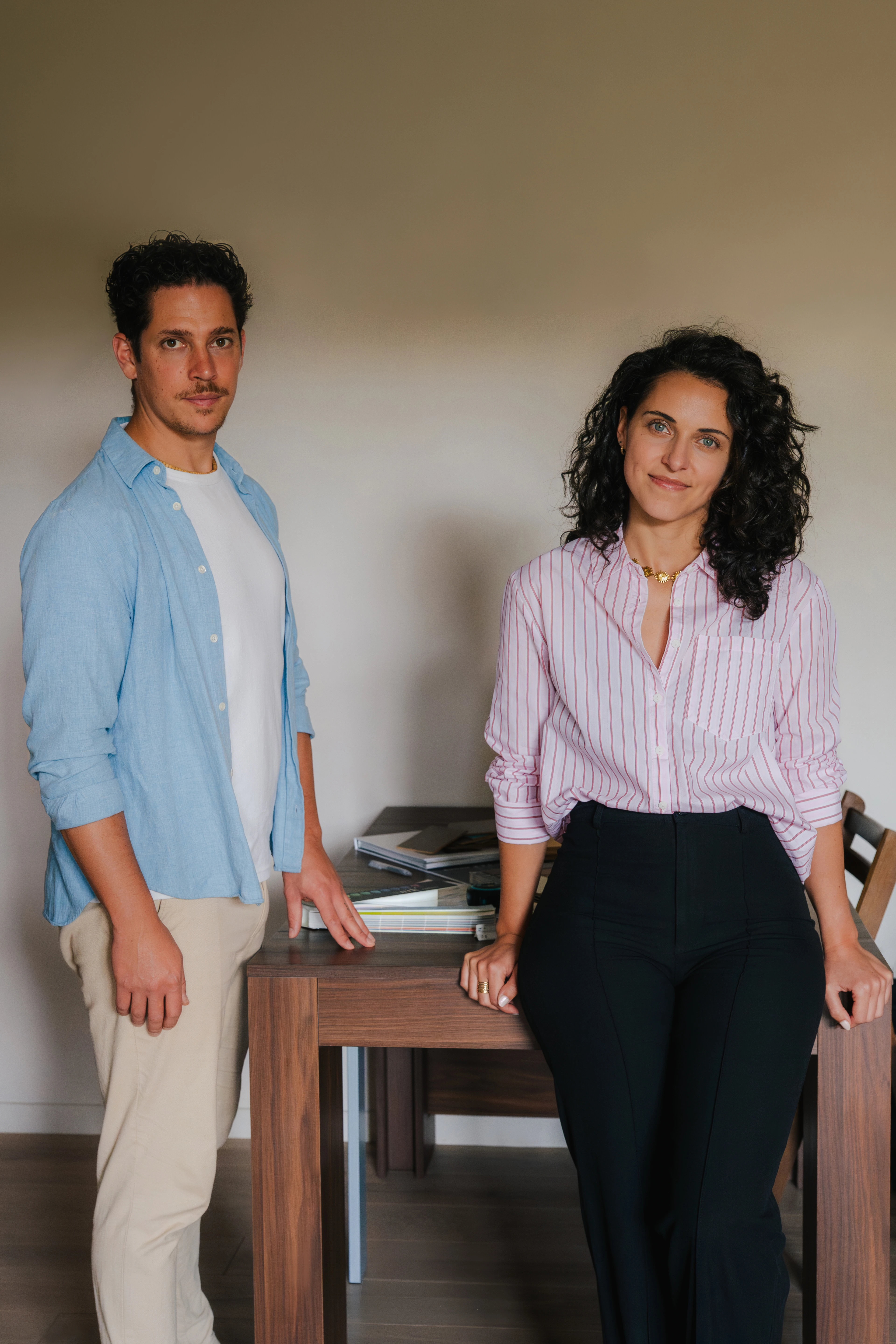 Deux professionnels souriants, un homme et une femme, debout dos à dos dans un bureau moderne avec des collègues travaillant en arrière-plan.