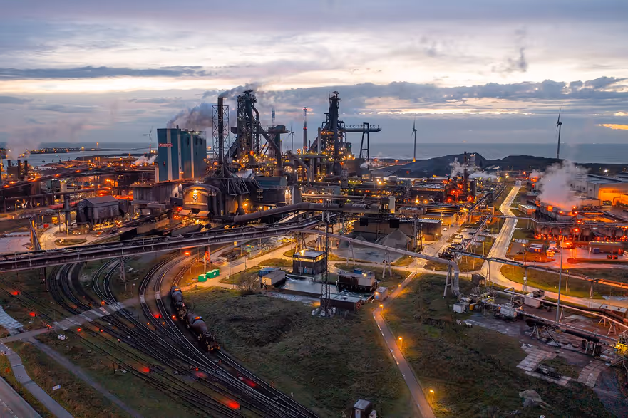 Industrieterrein bij zonsondergang met hoogovens, spoorlijnen en windturbines aan zee.