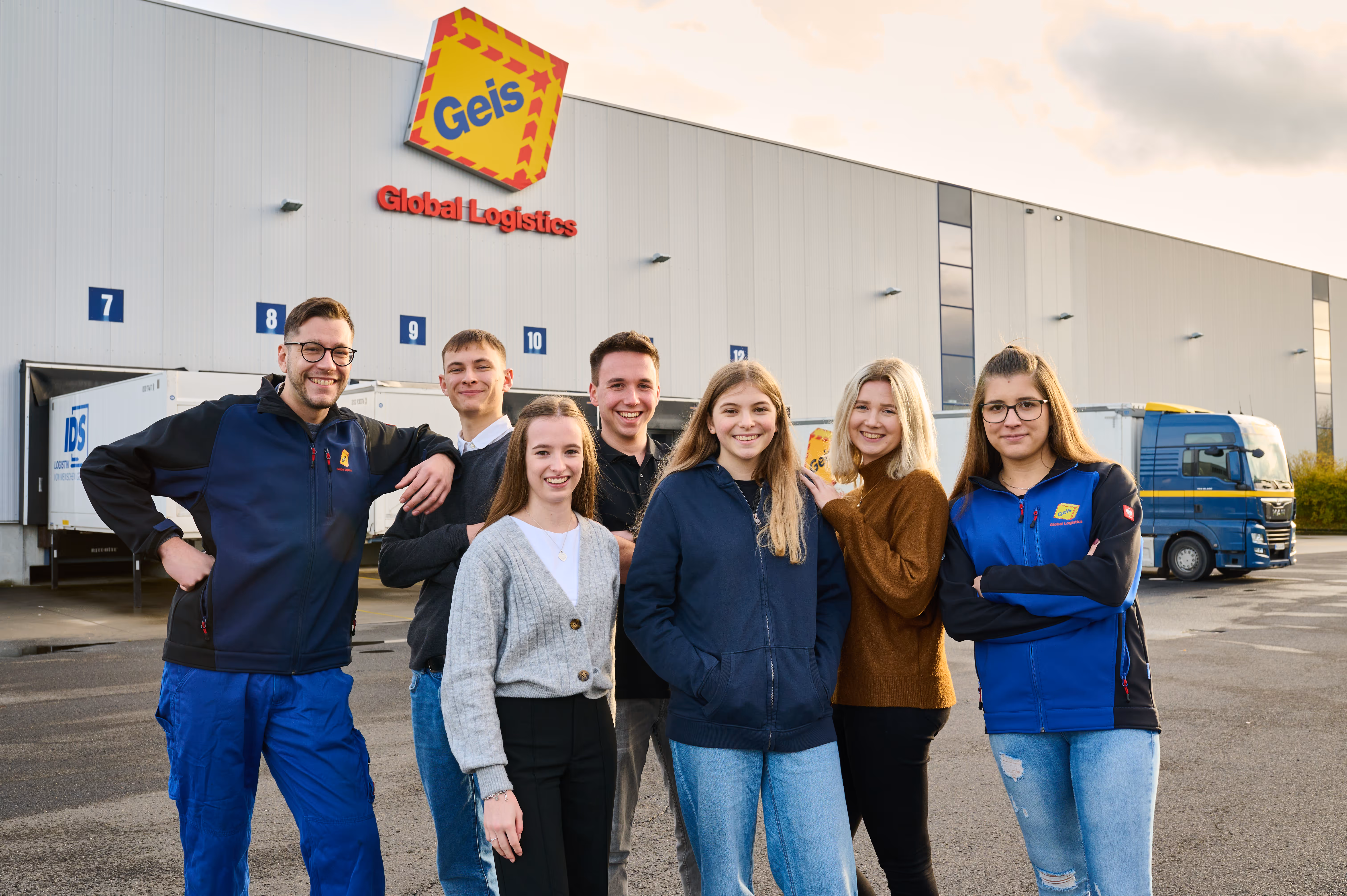 Sechs junge Mitarbeiter stehen lachend vor einem Logistikzentrum der Firma Geis mit Lastwagen im Hintergrund.
