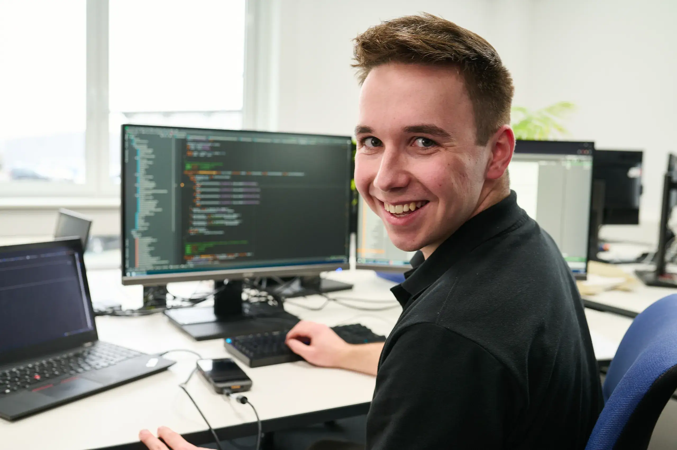Junger Mann sitzt am Arbeitsplatz vor mehreren Bildschirmen mit Programmiercode und schaut lächelnd zur Kamera.