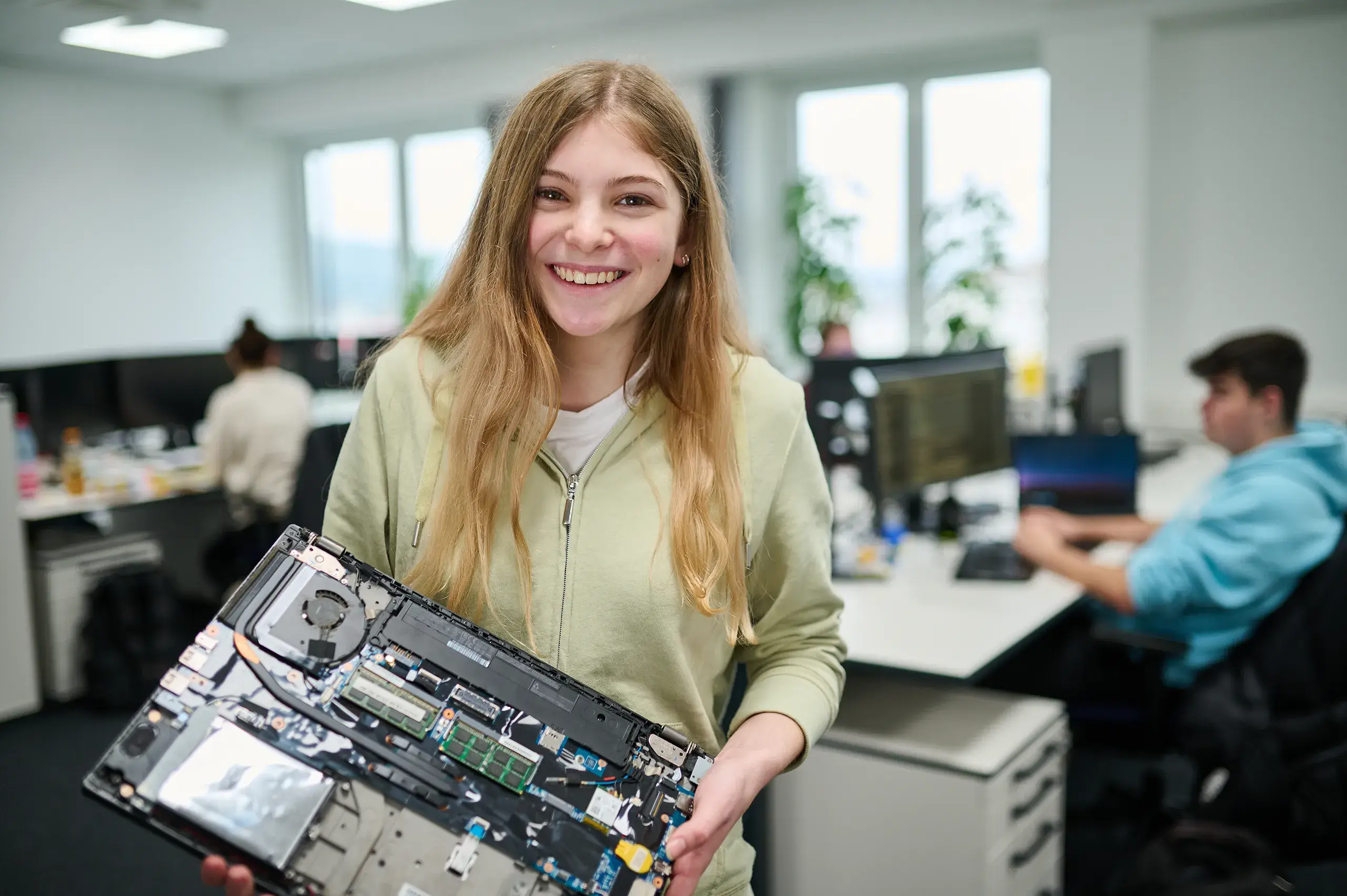 Junge Frau lächelt und hält offen gelegtes Laptop-Mainboard in einem modernen Büro mit zwei Personen am Computer im Hintergrund.