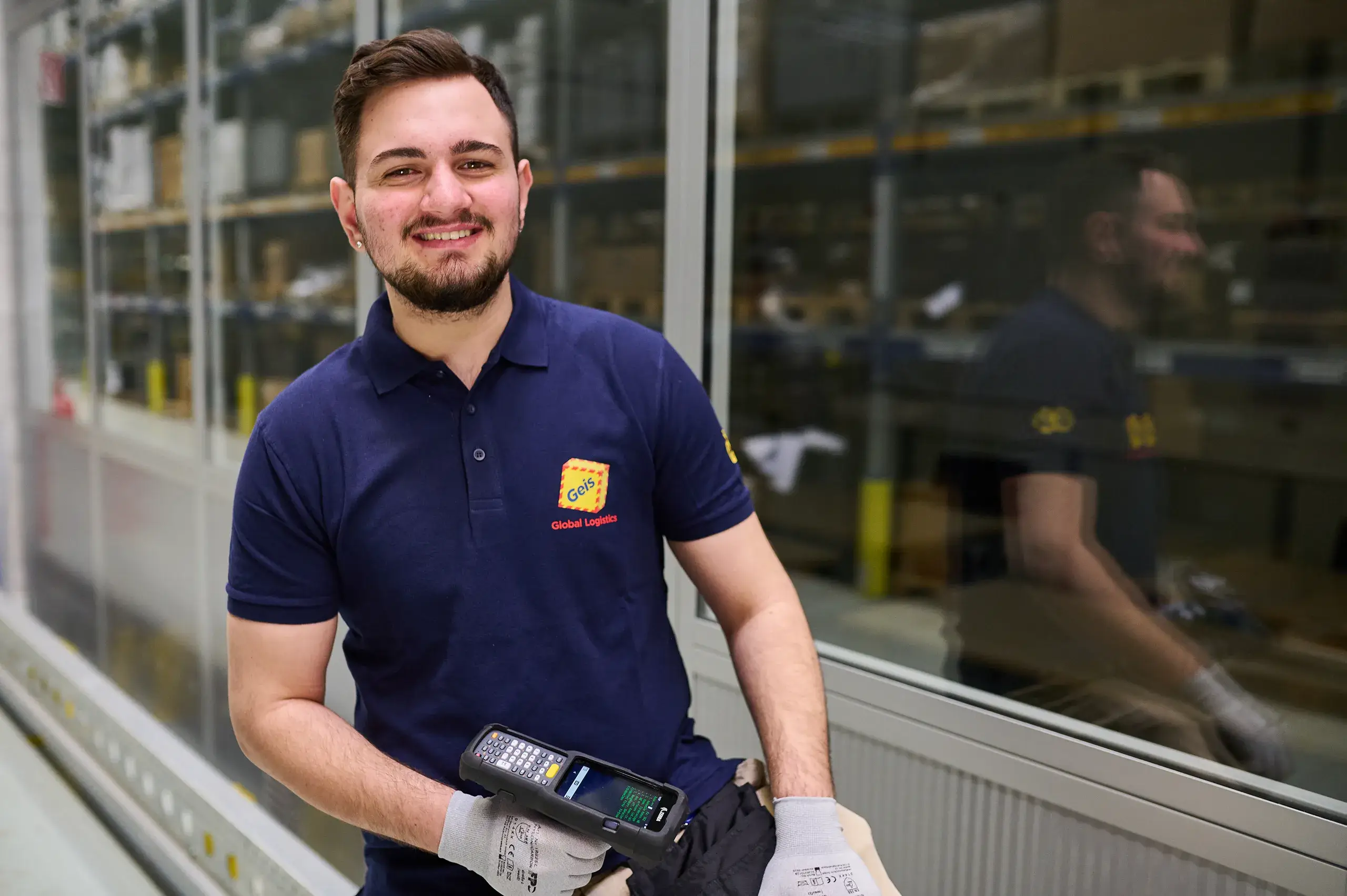 Junger Mann mit Geis Global Logistics Polohemd und Handschuhen hält ein mobiles Barcode-Scanner-Gerät in einem Lager.