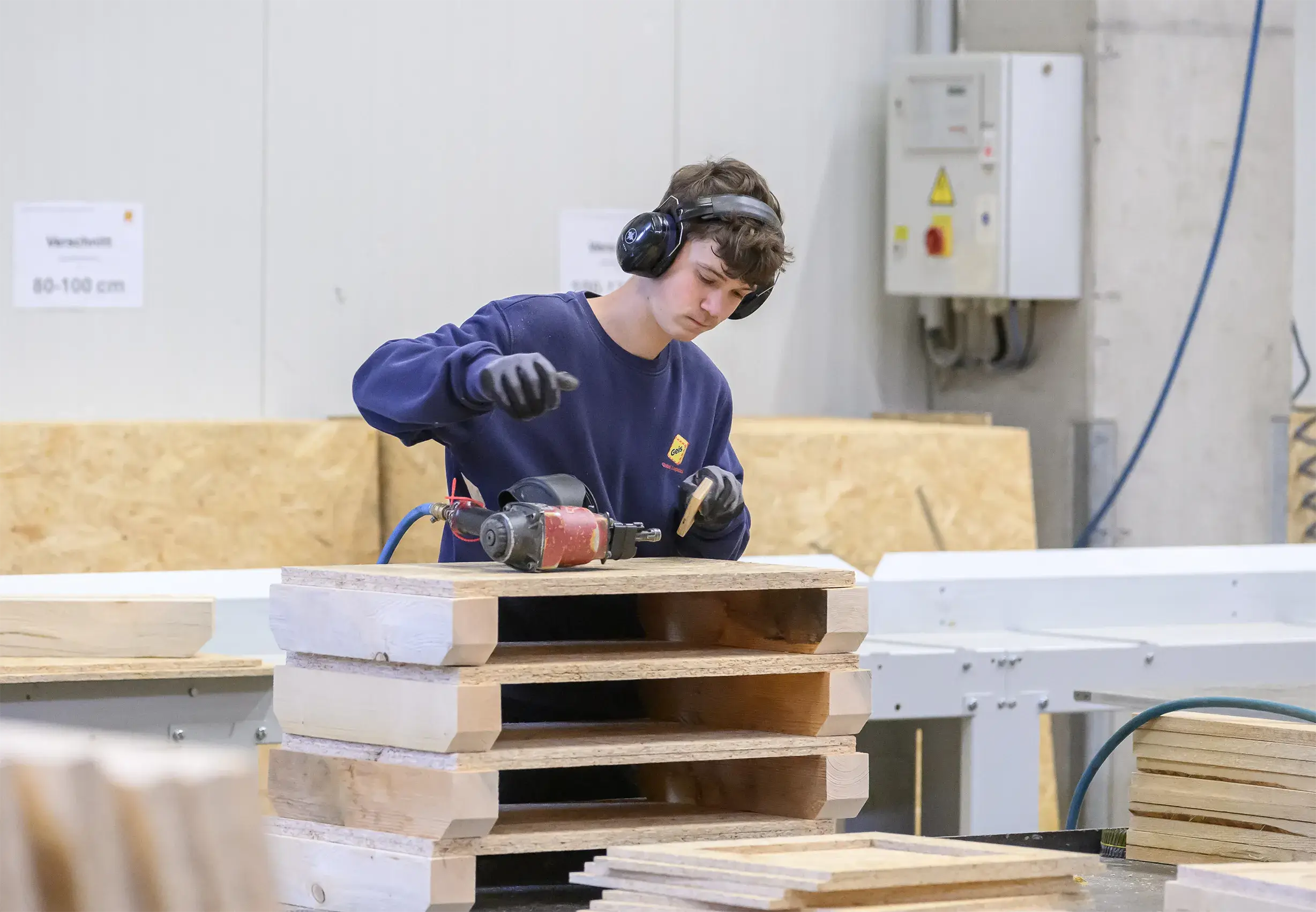 Junger Mann mit Gehörschutz und Handschuhen arbeitet mit einem Nagler an Holzpaletten in einer Werkstatt.