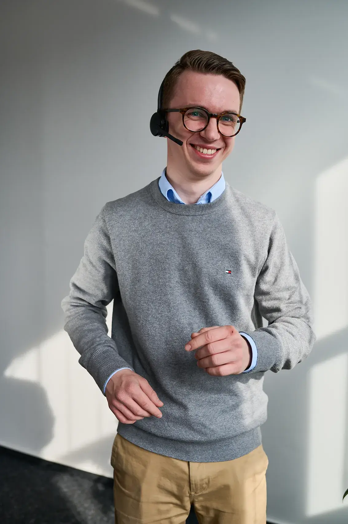 Junger Mann mit Brille, grauem Pullover und Headset lächelt freundlich.