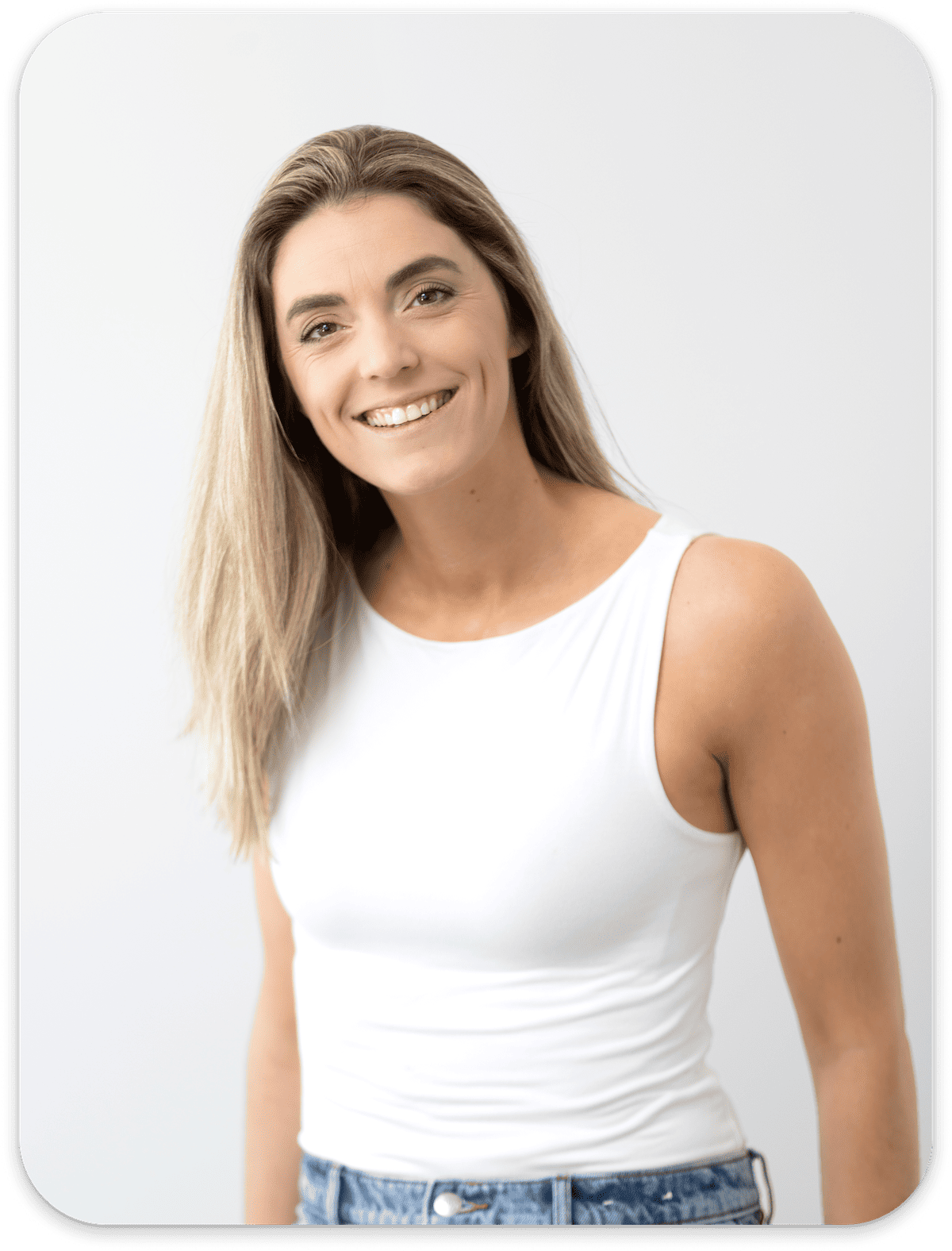 Smiling woman with long hair wearing a white sleeveless top and jeans against a plain background.
