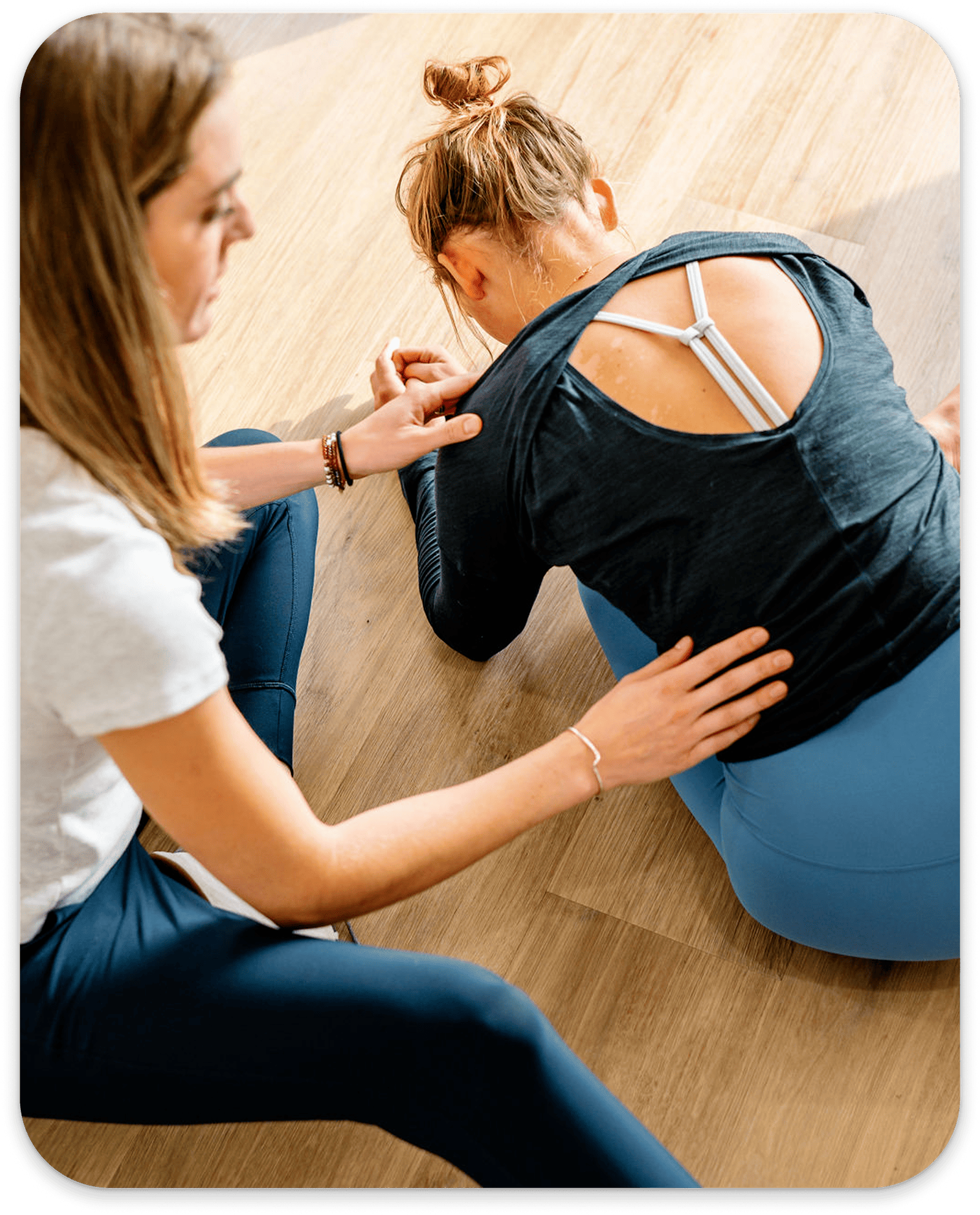 Physical therapist assisting woman in seated forward stretch on wooden floor.