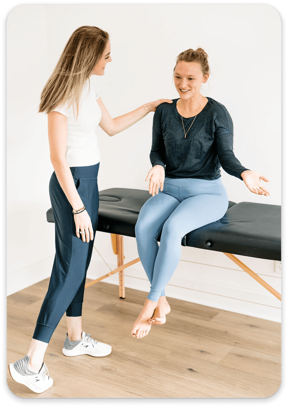 Physical therapist talks and comforts a barefoot woman sitting on an exam table in a bright room.