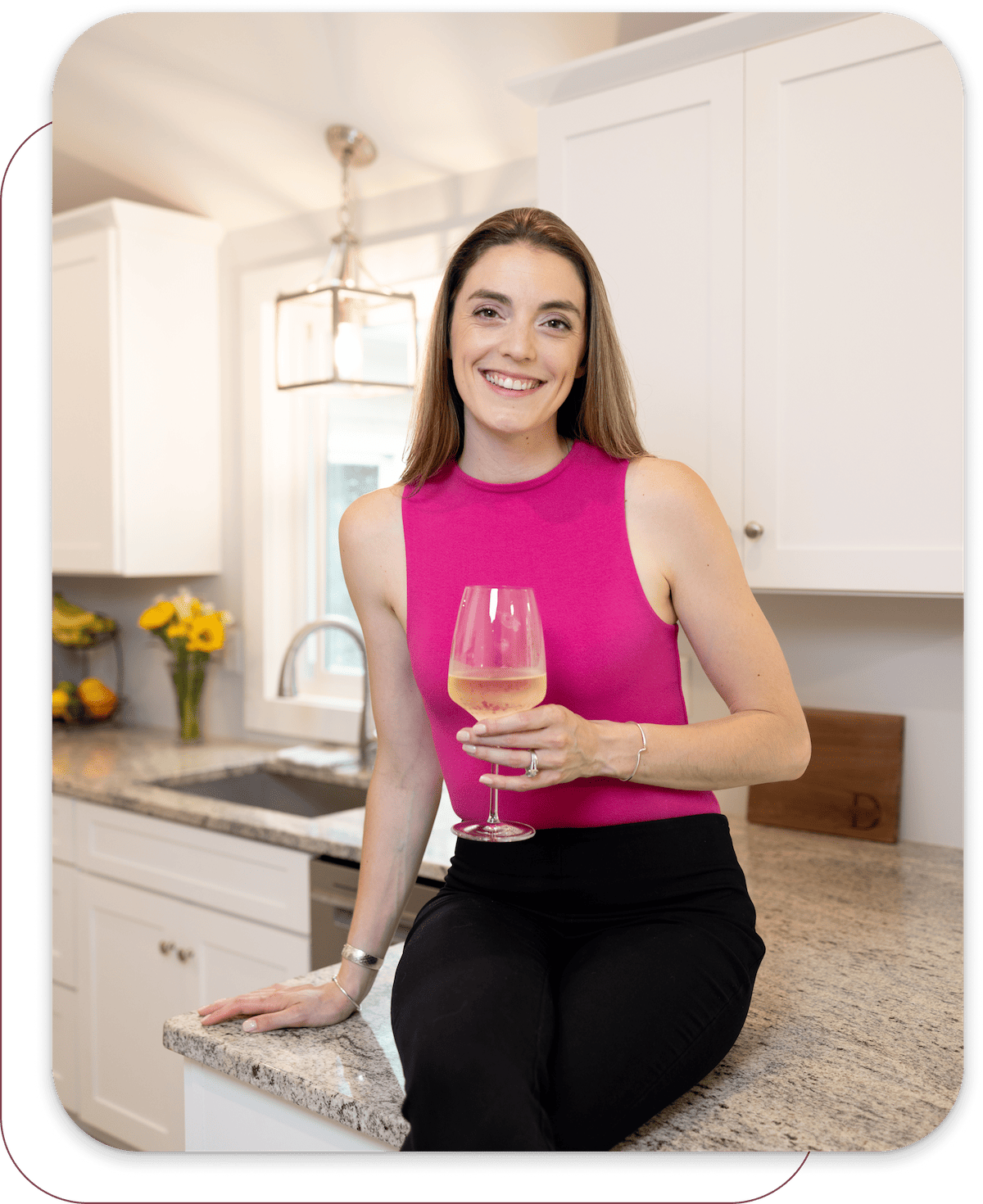 Smiling woman in a pink sleeveless top sitting on a kitchen counter holding a glass of white wine.