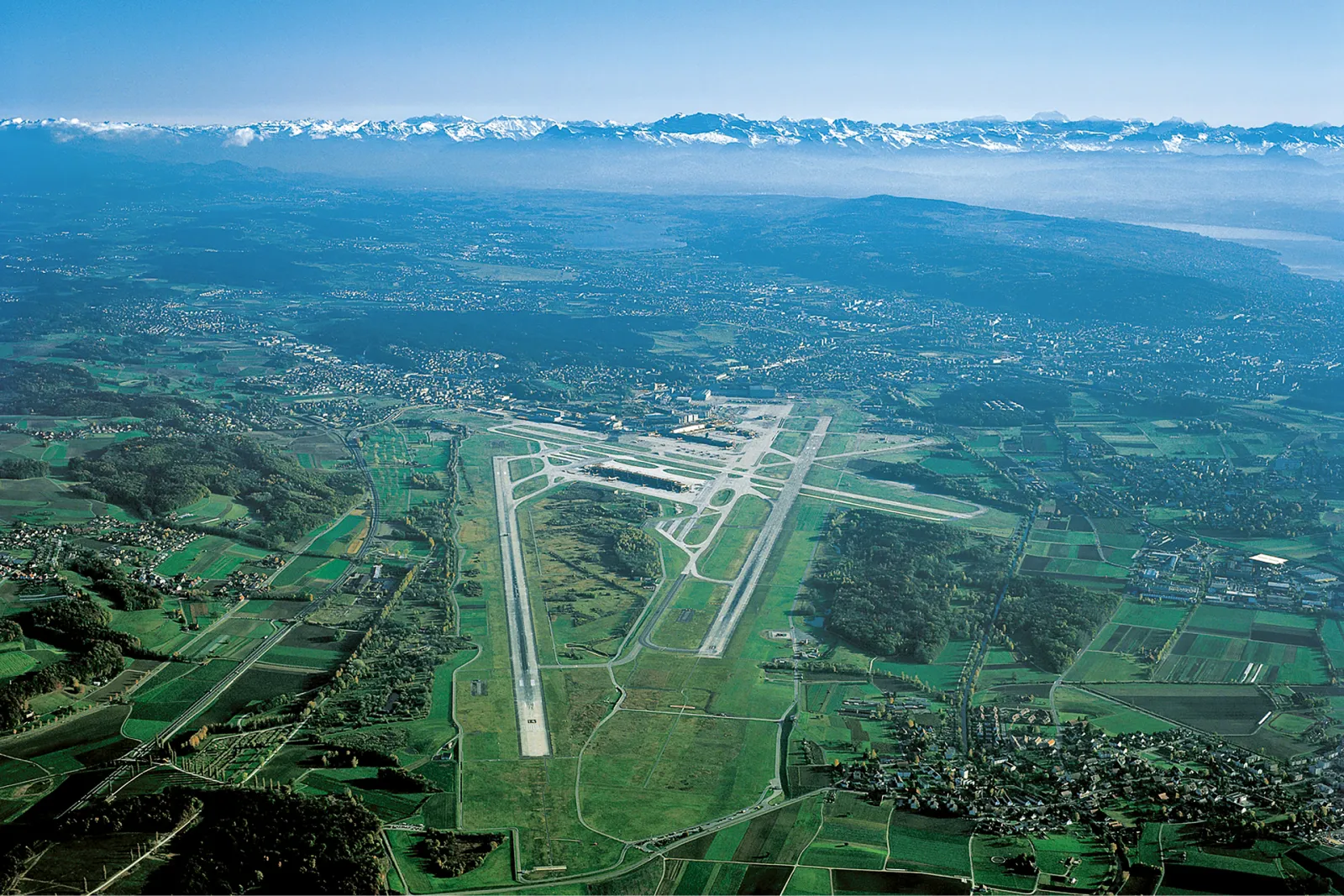 Luftaufnahme des Flughafens Zürich mit Start- und Landebahnen, umgeben von grünen Feldern und Bergen im Hintergrund.