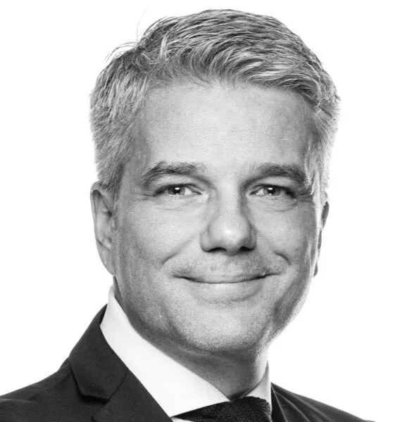 Porträt eines lächelnden Mannes mit grau meliertem Haar in Anzug und Krawatte vor weissem Hintergrund.